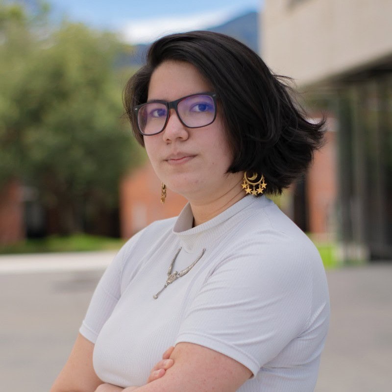 A person with short dark hair, glasses, and gold earrings stands outdoors with arms crossed, wearing a white top. Blurred buildings and greenery are in the background. | Wonderly
