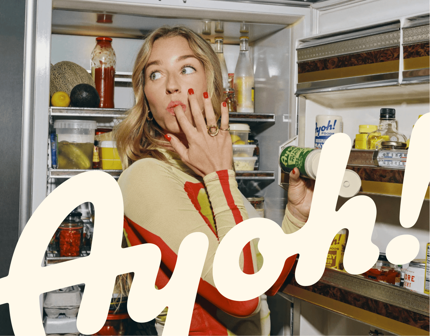 A woman stands in front of an open refrigerator, holding a jar and touching her lips, with the word “Ayoh!” in large text across the image. | Wonderly