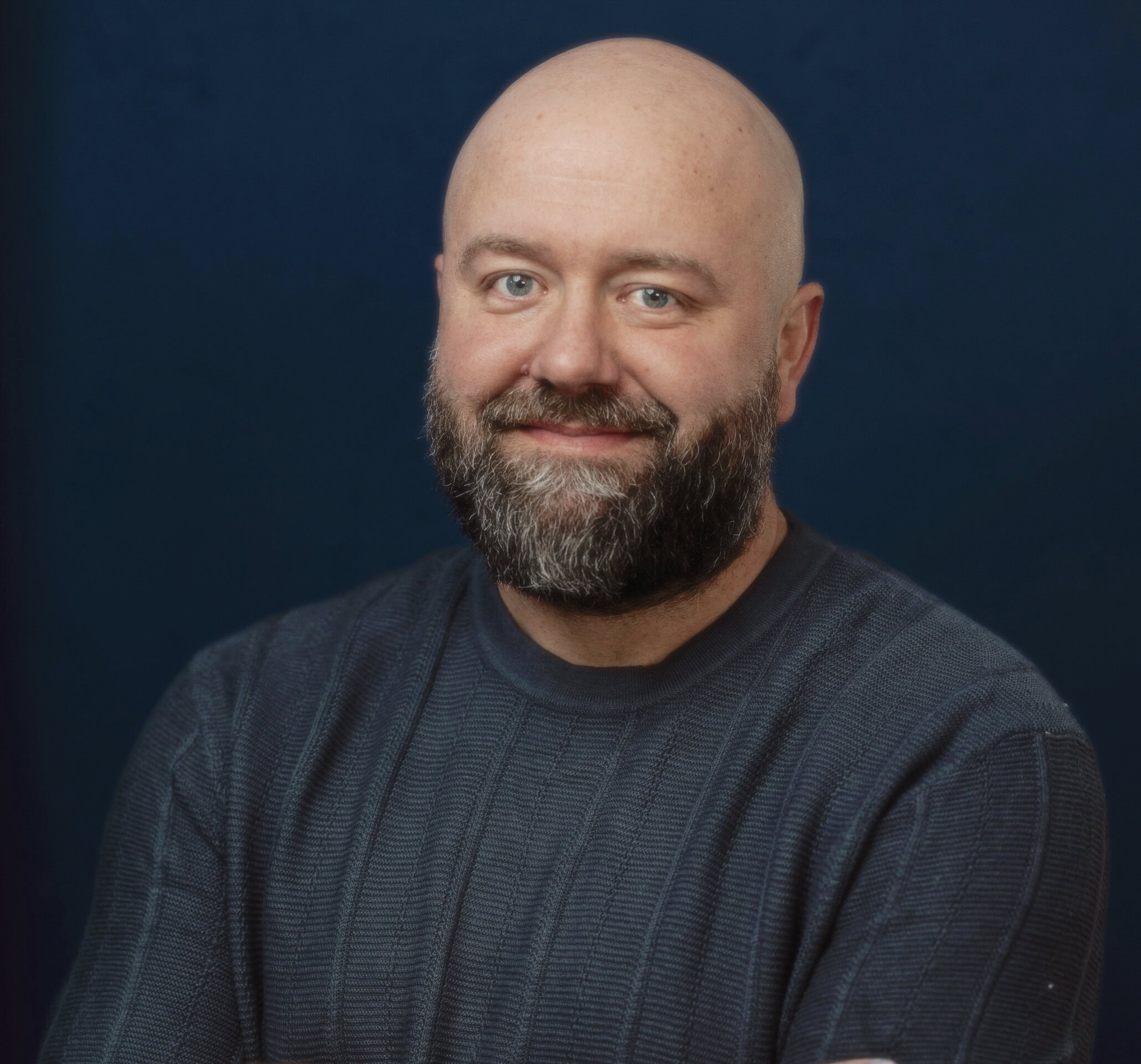 A bald man with a beard wearing a dark textured sweater stands against a dark blue background, smiling with arms crossed. | Wonderly