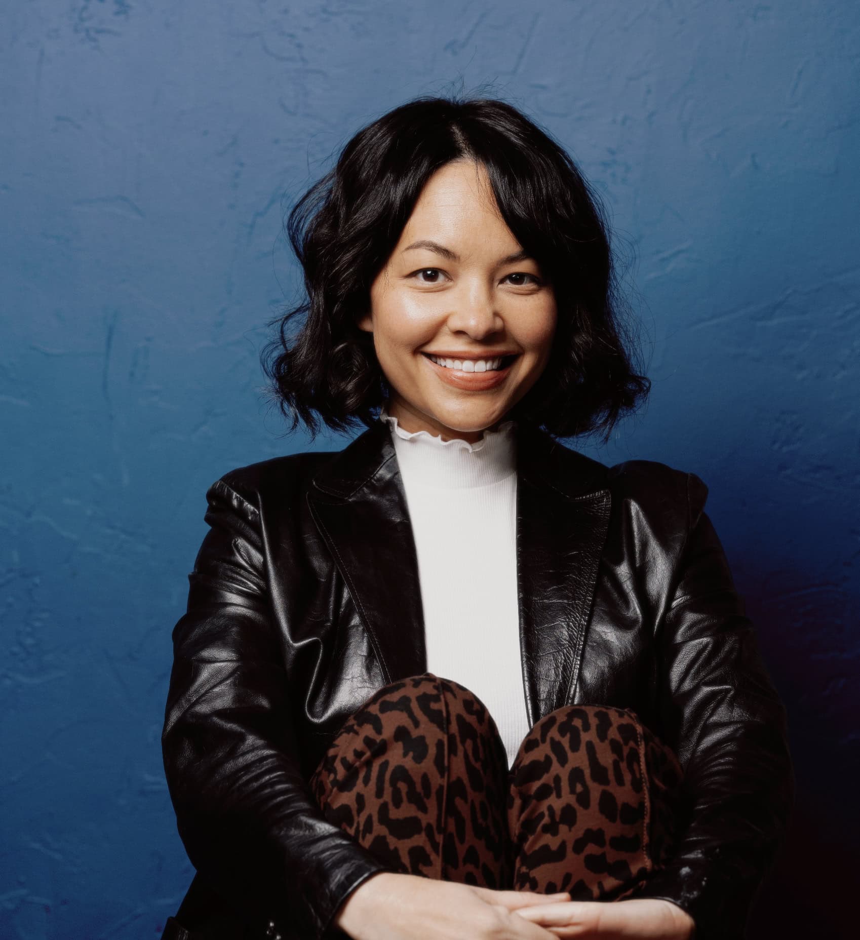 Woman with shoulder-length black hair sits against a textured blue wall, wearing a black leather jacket, white turtleneck, and leopard print pants, smiling at the camera. | Wonderly