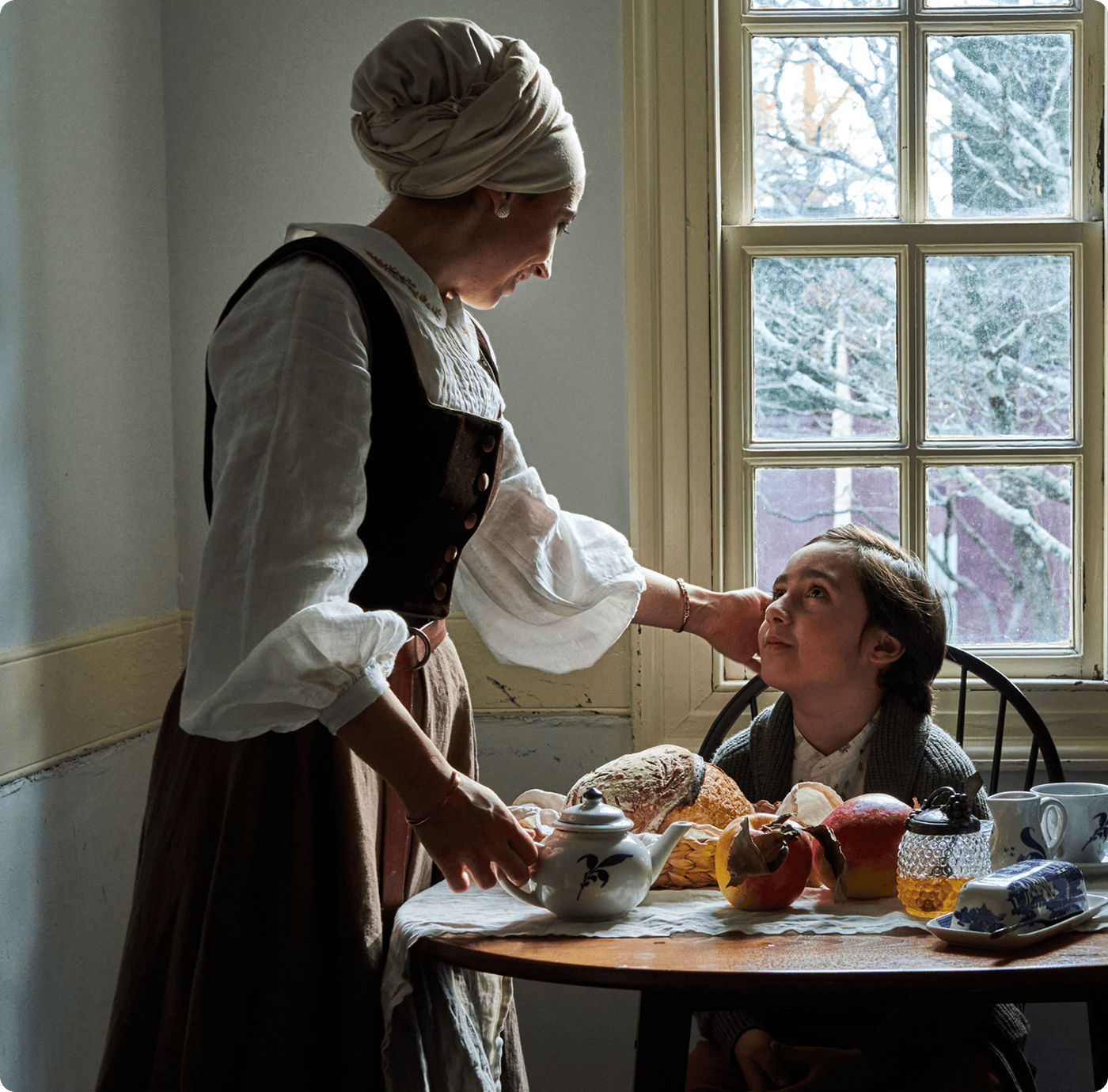 A woman in historical clothing serves tea and food to a seated girl at a small table by a window, with bread, fruit, and jam visible. | Wonderly