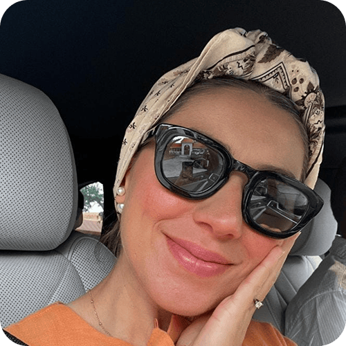 Woman wearing black sunglasses and a patterned headscarf, smiling and resting her face on her hand while sitting in a car. | Wonderly