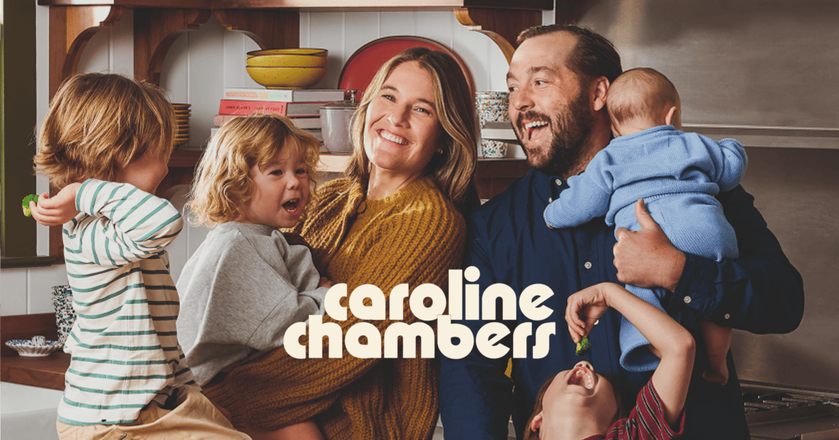 A smiling couple stands in a kitchen holding three young children. "Caroline Chambers" text is overlaid in bold letters at the bottom. | Wonderly