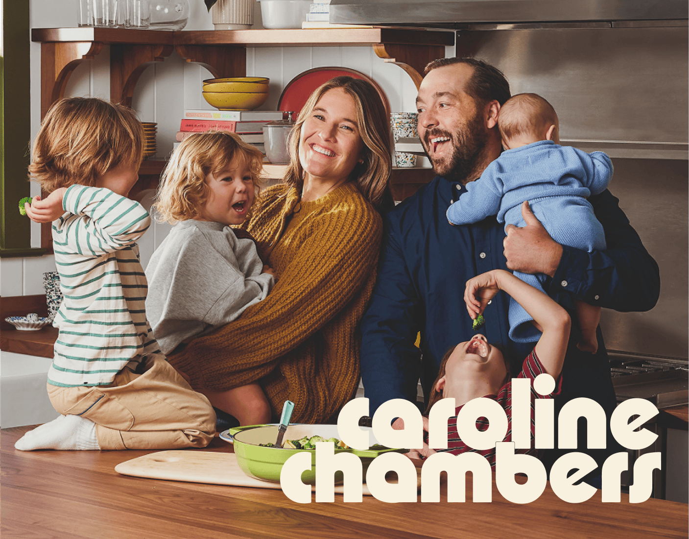 A smiling family of five gathers in a kitchen, with two adults holding three young children. The text "caroline chambers" is displayed in bold letters at the bottom right. | Wonderly