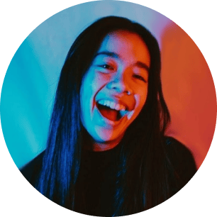 A person with long hair smiles broadly, lit by colorful blue and red lighting. | Wonderly