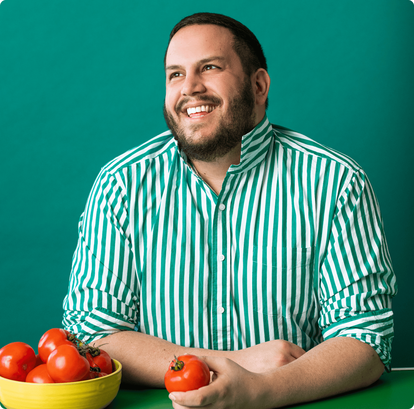 A person in a green and white striped shirt sits at a table with a bowl of tomatoes, holding one tomato and smiling against a green background. | Wonderly