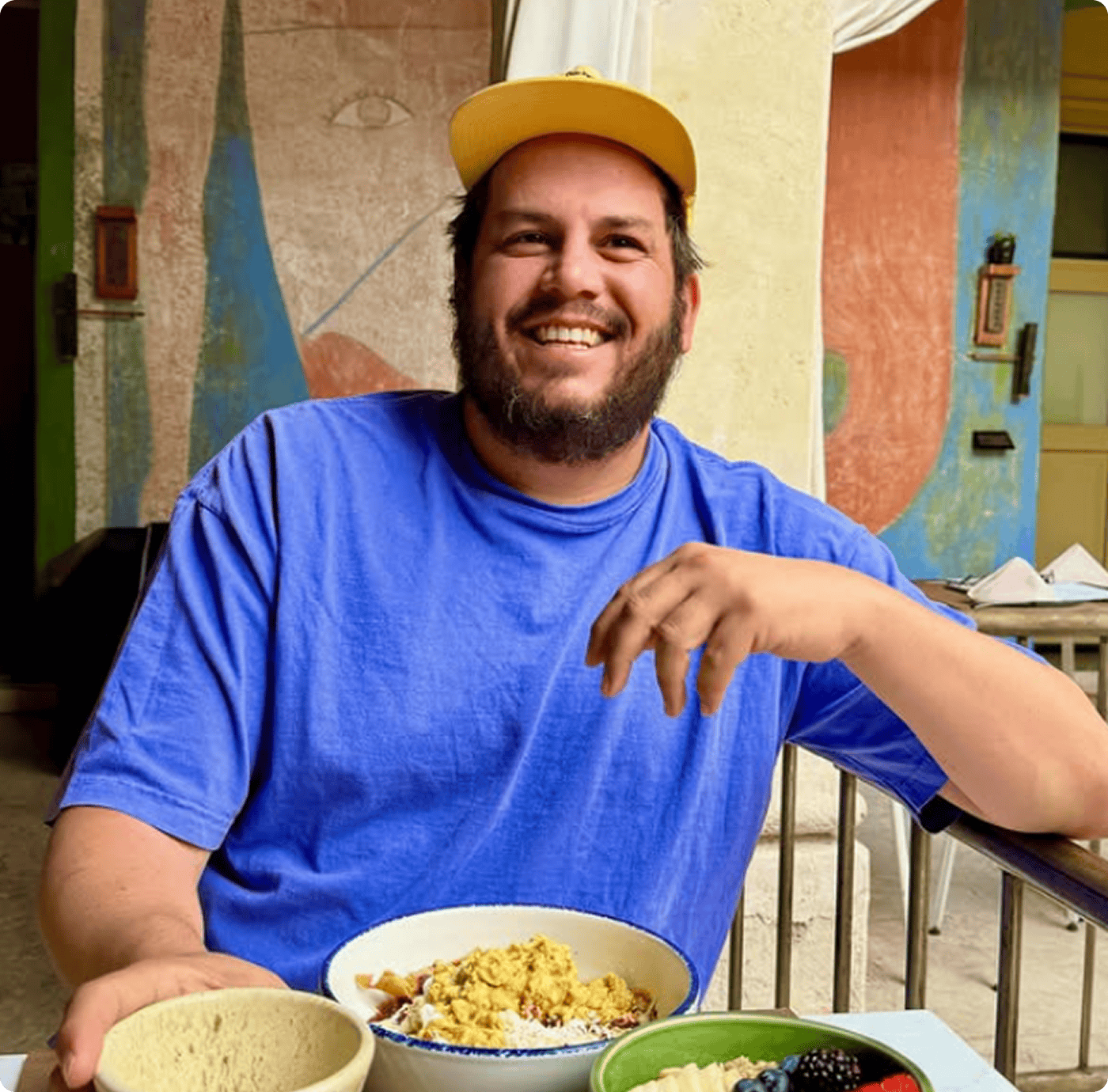 A man wearing a yellow cap and blue shirt sits at a table with bowls of food, smiling at the camera in an outdoor setting with artistic murals in the background. | Wonderly