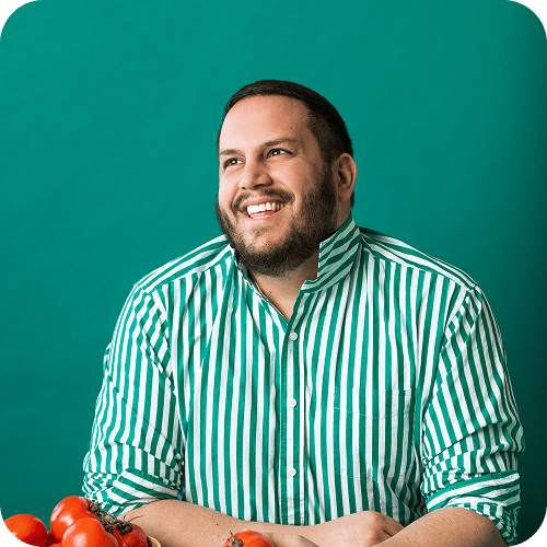 A man in a green and white striped shirt sits at a table with tomatoes, smiling and looking slightly upward. The background is solid teal. | Wonderly