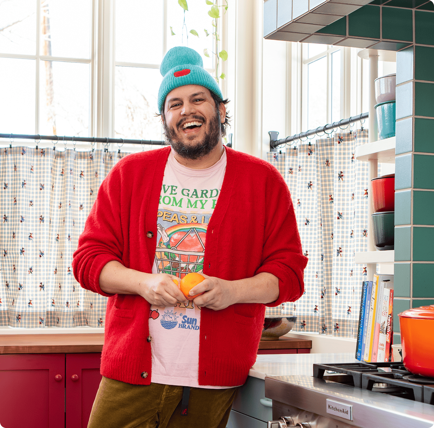 A person wearing a blue beanie, red cardigan, and graphic t-shirt stands in a colorful kitchen, smiling and holding an orange. | Wonderly