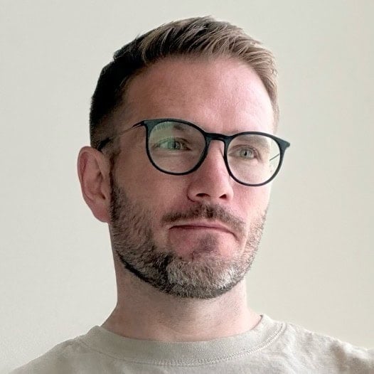 A man with short hair, a trimmed beard, and glasses looks to the side against a plain light background. | Wonderly