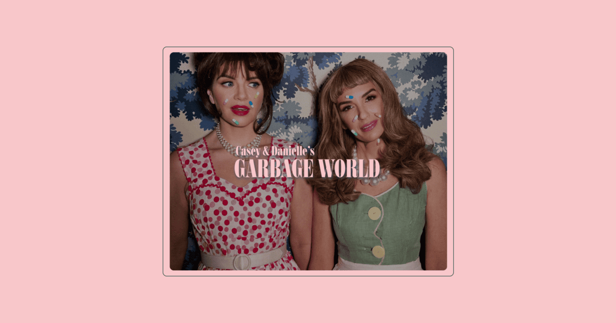 Two women in vintage-style dresses stand side by side against a floral wallpaper background. Text reads, "Casey & Danielle's Garbage World. | Wonderly