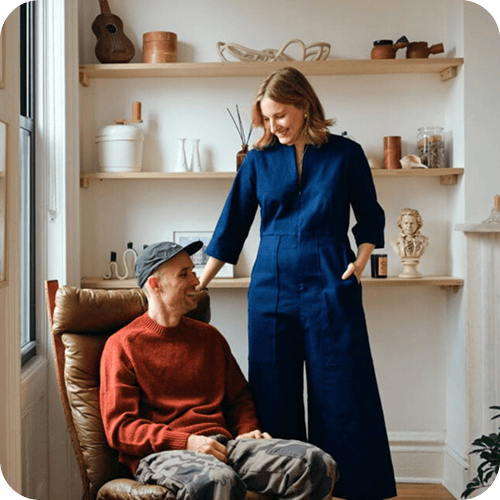 A man sits in a brown chair while a woman in a blue jumpsuit stands beside him in a room with wooden shelves and various decorative items. | Wonderly