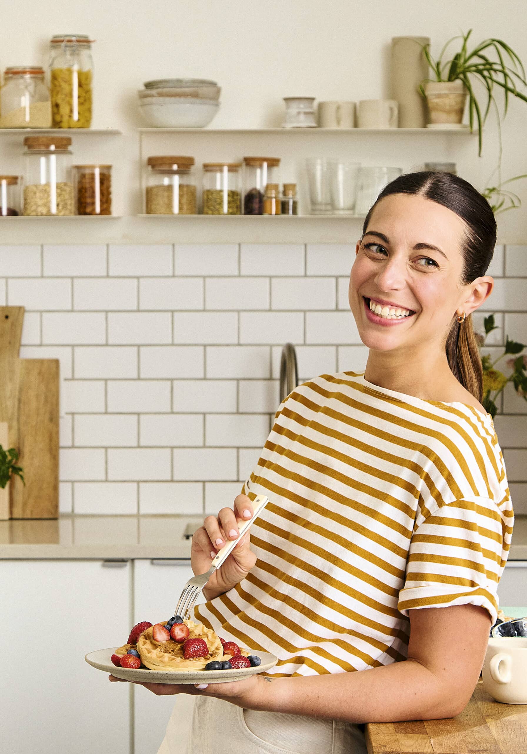 Woman in a striped shirt smiles while holding a plate of pancakes topped with berries in a modern kitchen. | Wonderly