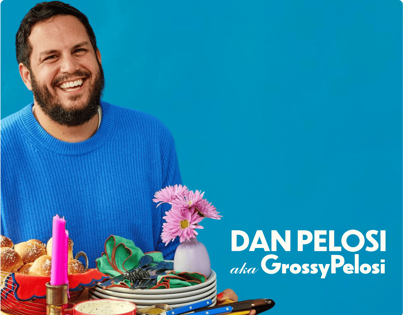 A smiling man in a blue sweater sits at a table with colorful dishes, candles, and a vase of pink flowers. Text reads "Dan Pelosi aka GrossyPelosi" on a blue background. | Wonderly