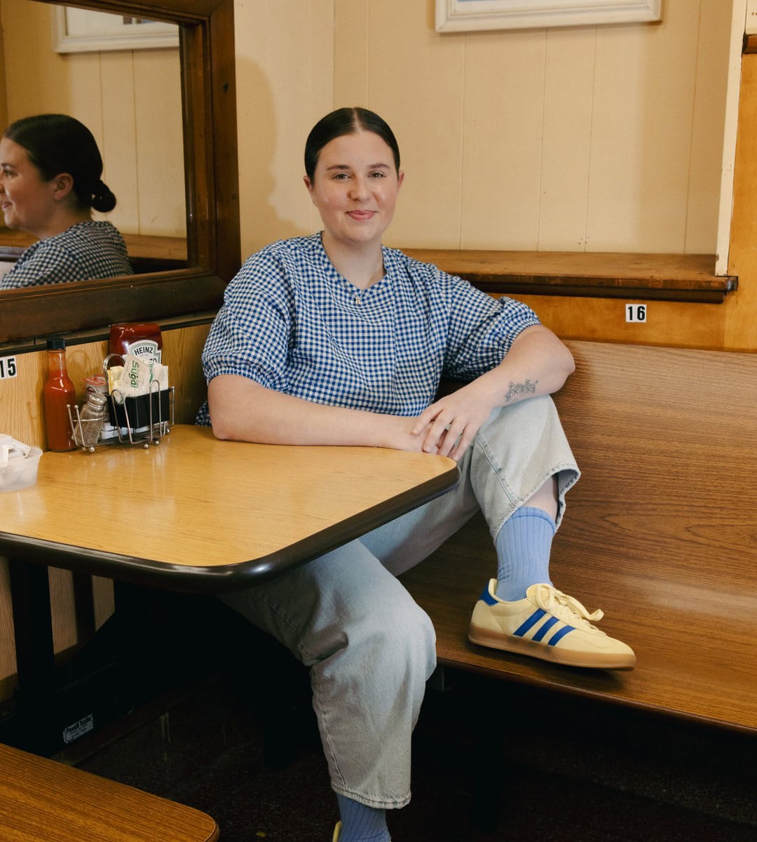 A person in a blue checkered shirt, light jeans, and yellow Adidas sneakers sits in a wooden booth at a diner, with condiments on the table and their reflection visible in the mirror. | Wonderly