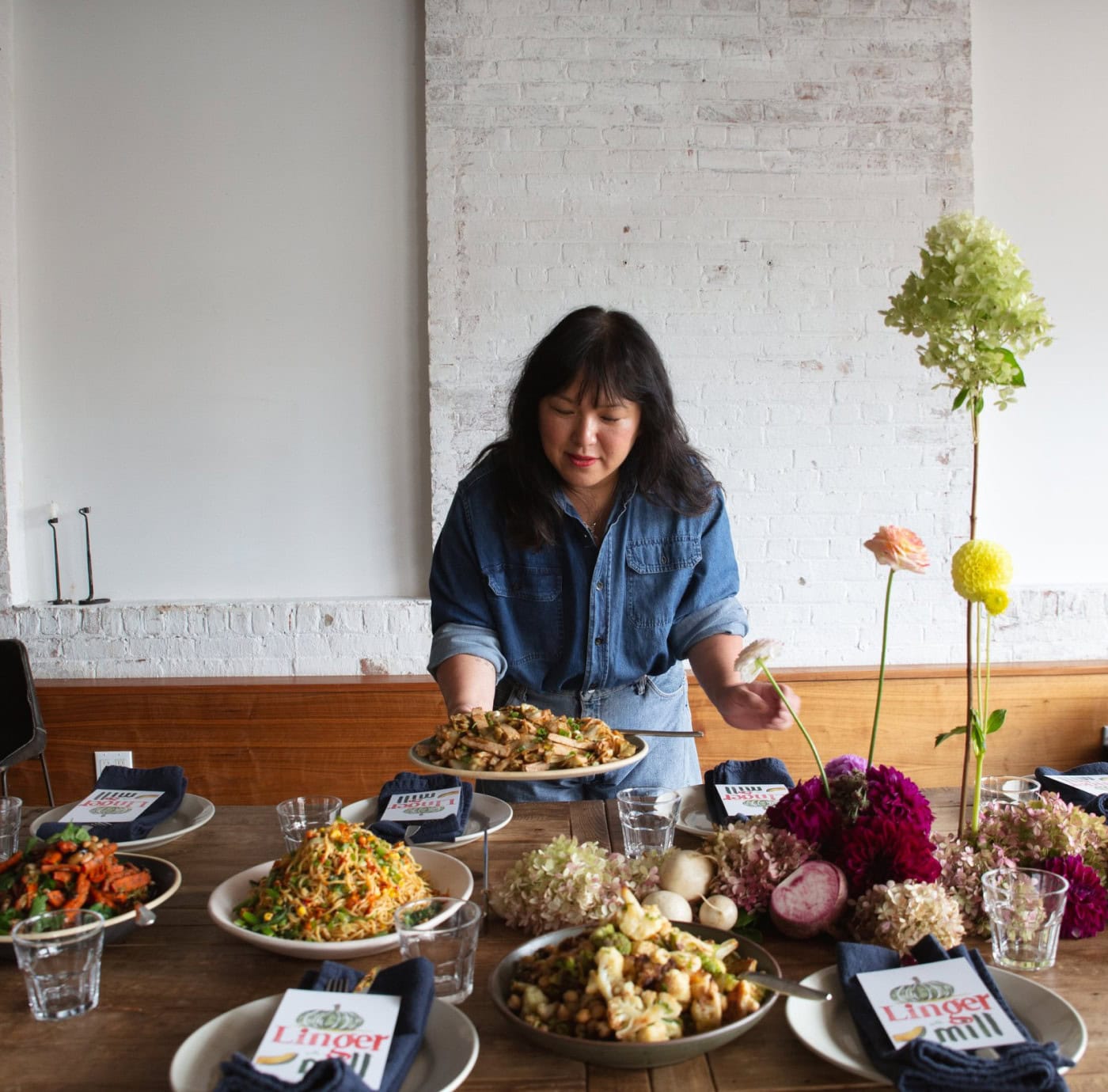 A woman in a denim shirt sets dishes of food on a wooden table arranged with napkins, menus, flowers, and various plates of prepared meals in a bright room with a white brick wall. | Wonderly