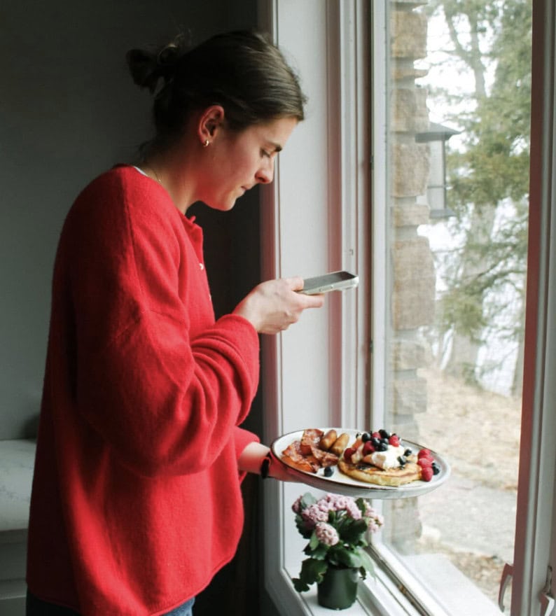 A person in a red sweater stands by a window, holding a plate of waffles topped with fruit and using a smartphone to take a photo of it. | Wonderly