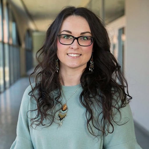 A woman with long curly hair, glasses, and a blue top stands in a bright hallway, looking at the camera and smiling. | Wonderly
