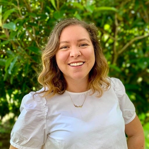 A woman with wavy hair wearing a white shirt smiles at the camera while standing outdoors in front of green foliage. | Wonderly