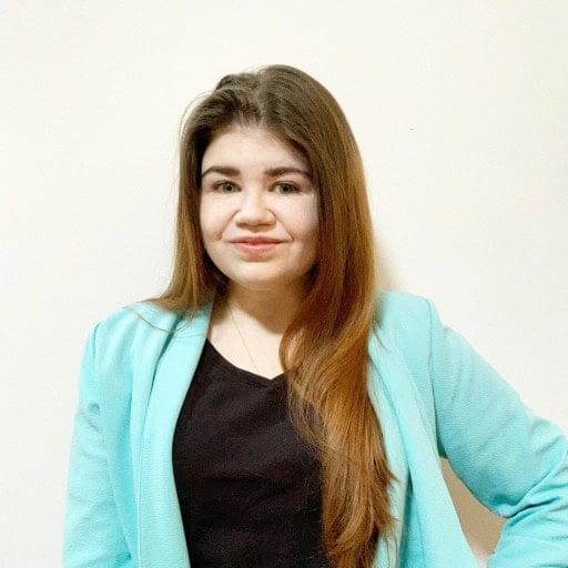 A woman with long brown hair wearing a light blue blazer over a black top stands against a plain light-colored wall, smiling slightly at the camera. | Wonderly