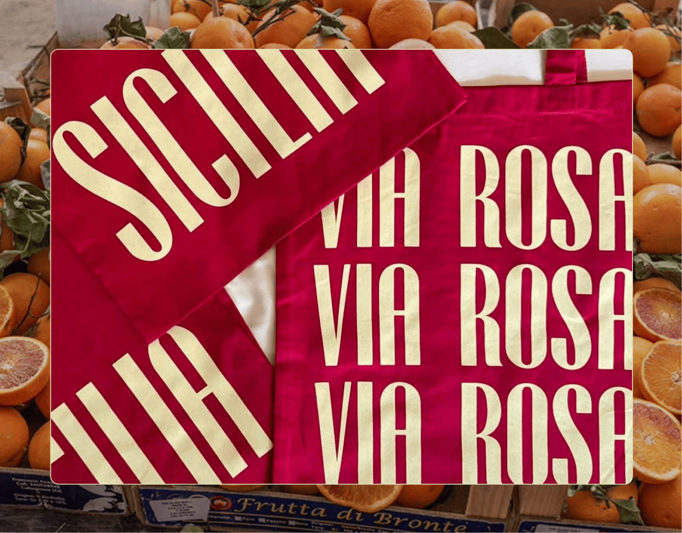 Red tote bags with bold, cream-colored text reading "SICILIA" and "VIA ROSA" displayed over a background of fresh oranges. | Wonderly