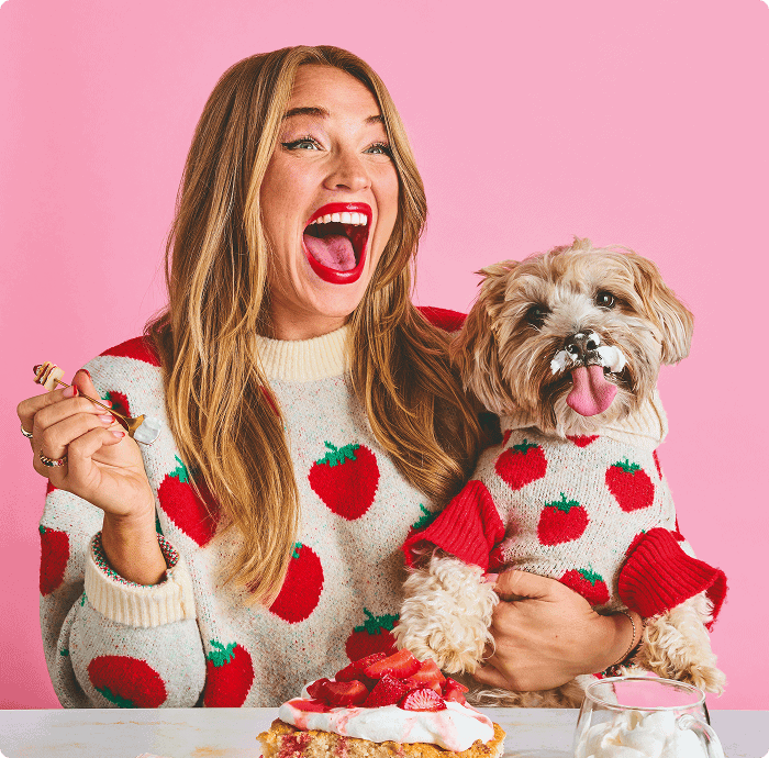 A woman and a small dog wearing matching strawberry sweaters sit in front of a strawberry dessert; the dog has whipped cream on its nose. | Wonderly