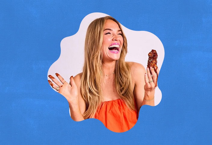 A woman with long blonde hair wearing an orange top laughs while holding a piece of food in one hand against a blue background. | Wonderly