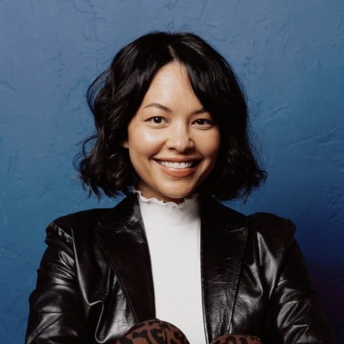 Woman with wavy black hair, wearing a black leather jacket and white top, smiling in front of a textured blue background. | Wonderly