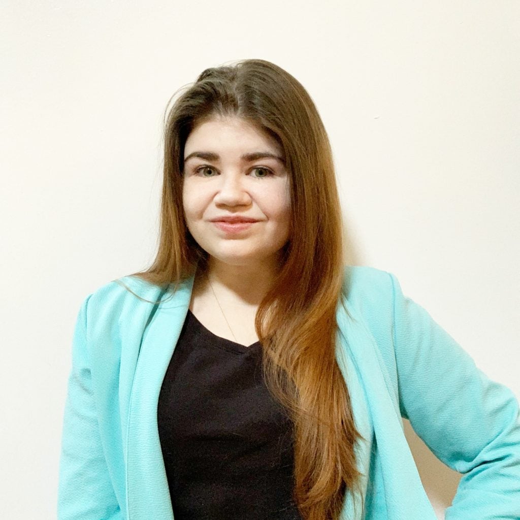 Woman with long brown hair wearing a turquoise blazer and black top stands in front of a plain white wall, looking at the camera with a slight smile. | Wonderly