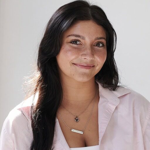 A person with long dark hair, in a light pink shirt, smiles at the camera. They wear layered necklaces. | Wonderly