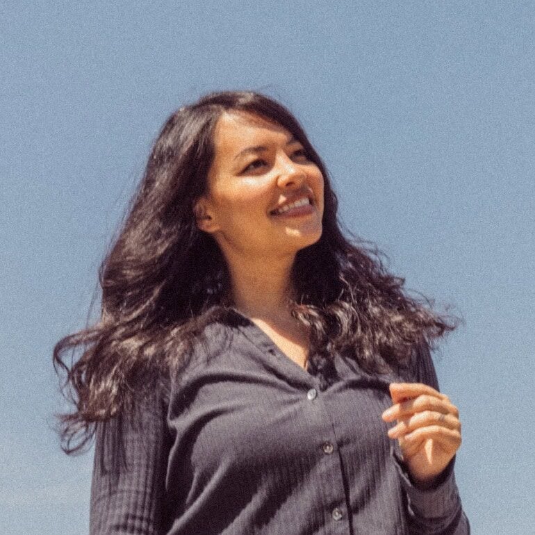 A woman with long dark hair wearing a dark button-up shirt stands outdoors, smiling and looking slightly to the side, with a clear blue sky in the background. | Wonderly