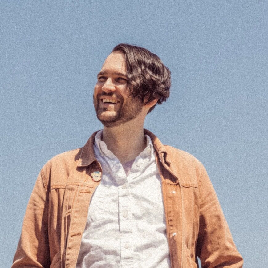 A man with wavy brown hair and a beard smiles while standing outside under a clear blue sky, wearing a light brown jacket over a white shirt. | Wonderly