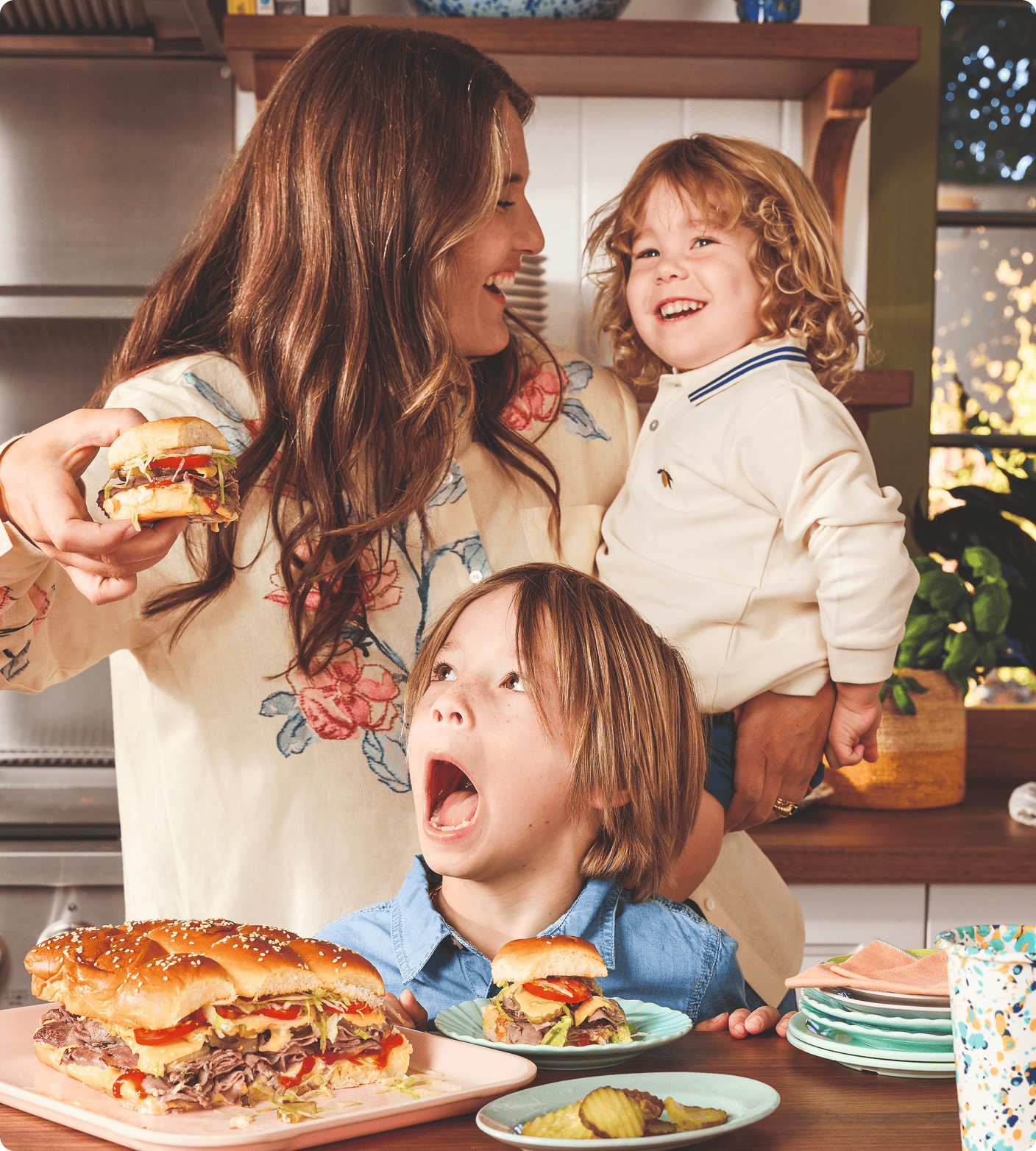 A woman holding a sandwich stands with two children in a kitchen, with sandwiches, pickles, and plates of food on the counter in front of them. | Wonderly