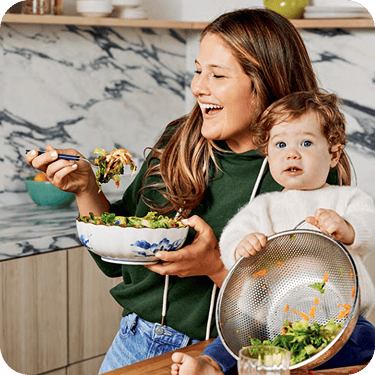 A woman smiles while eating salad, holding a bowl, as a toddler sits on the counter holding a colander with salad ingredients. | Wonderly
