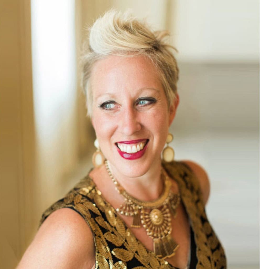 A woman with short blonde hair, red lipstick, and gold jewelry smiles while looking to the side in a well-lit indoor setting. | Wonderly