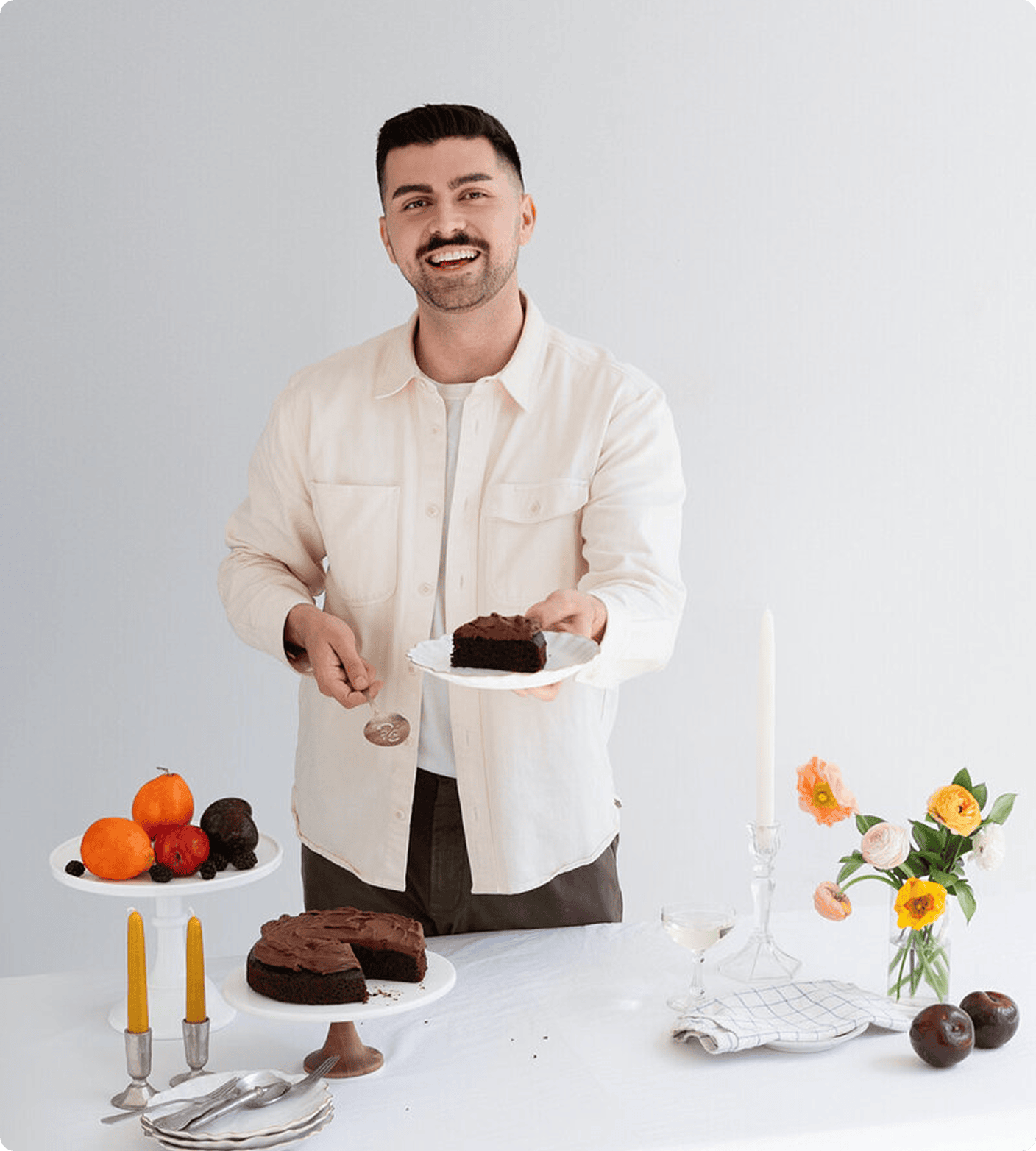 A man stands at a table holding a slice of chocolate cake. The table displays fruit, candles, flowers, utensils, and the rest of the cake. | Wonderly