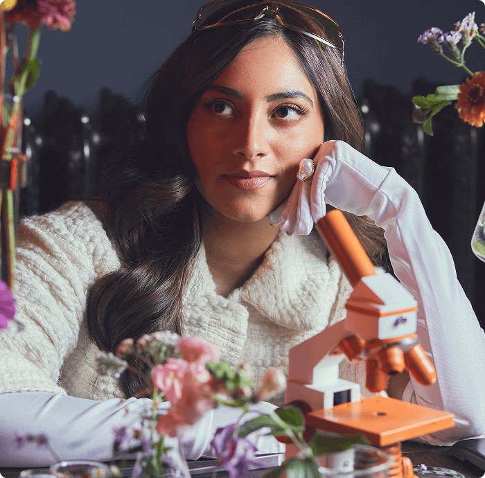 A woman in a white outfit sits at a table with a microscope and flowers, resting her chin on her hand and looking to the side. | Wonderly