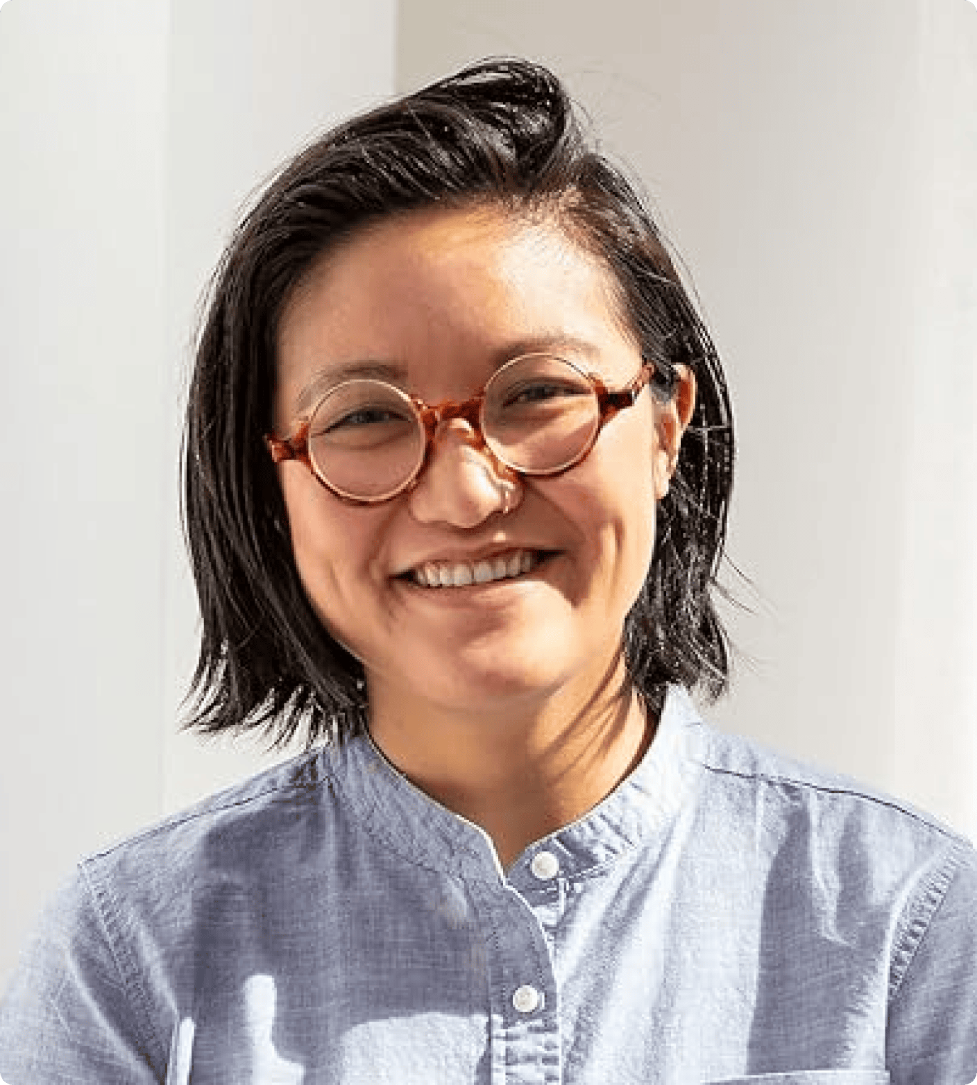 Person with short dark hair and round glasses, wearing a light blue button-up shirt, smiling in a brightly lit indoor setting. | Wonderly