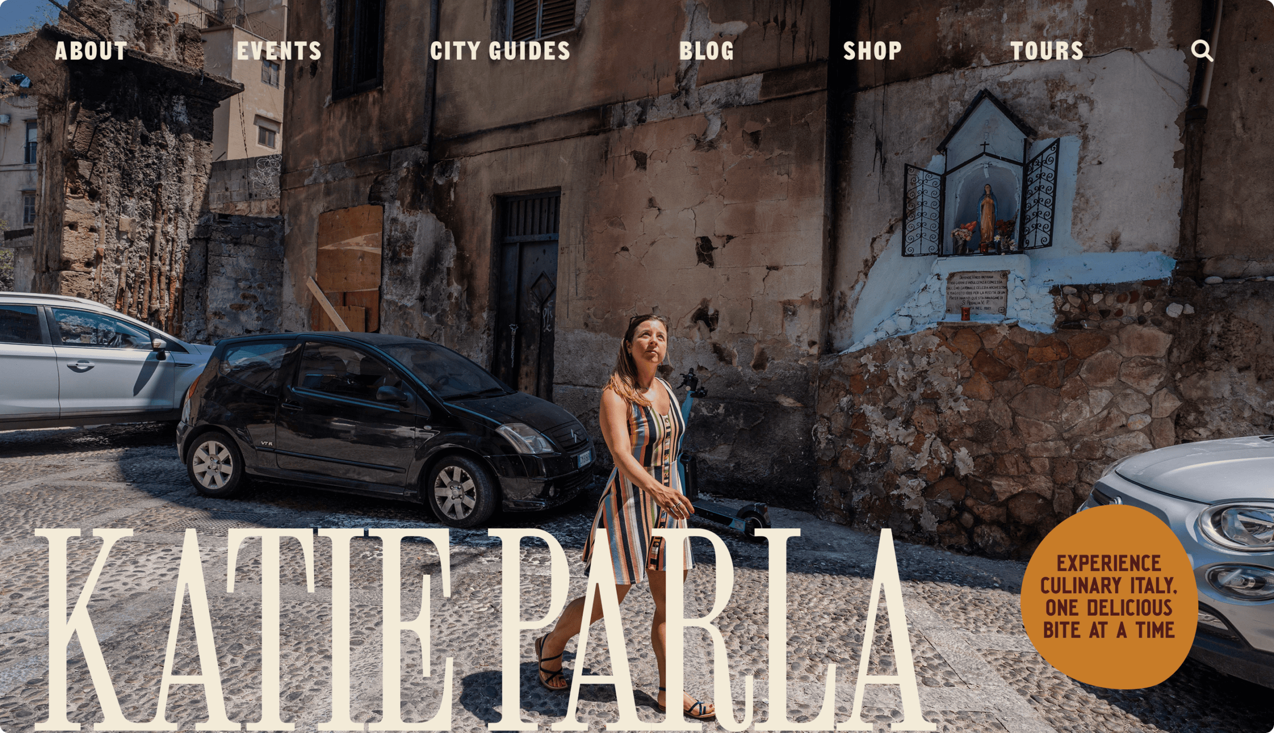 A woman walks on a cobblestone street beside parked cars and old buildings, with a small religious shrine set into the wall behind her. | Wonderly