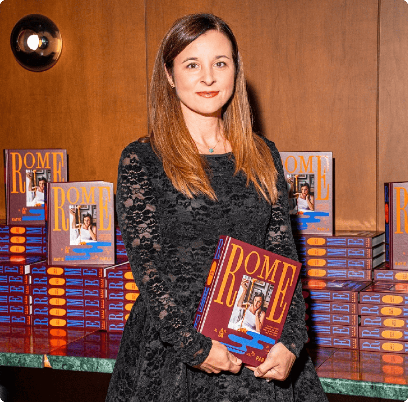 A woman in a black lace dress stands holding a book titled "ROME" in front of a display of stacked copies of the same book against a wooden wall. | Wonderly