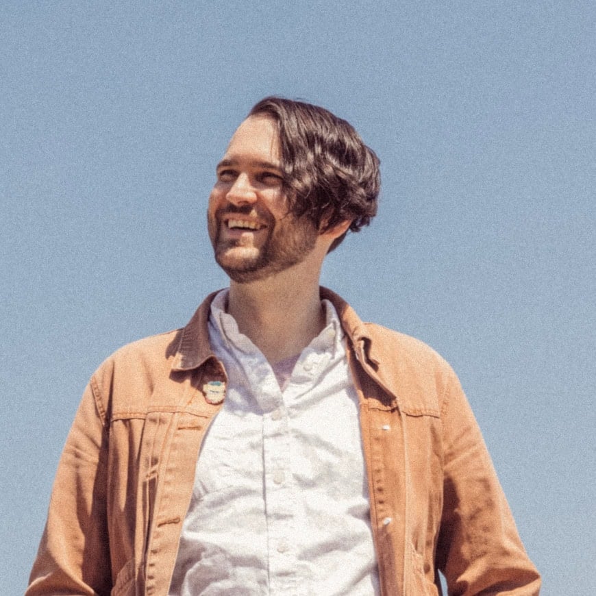 A man with wavy brown hair and a beard smiles while standing outside under a clear blue sky, wearing a light brown jacket over a white shirt. | Wonderly